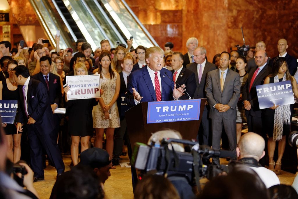 Trump at a podium addressing cameras with a crowd of supporters behind him.