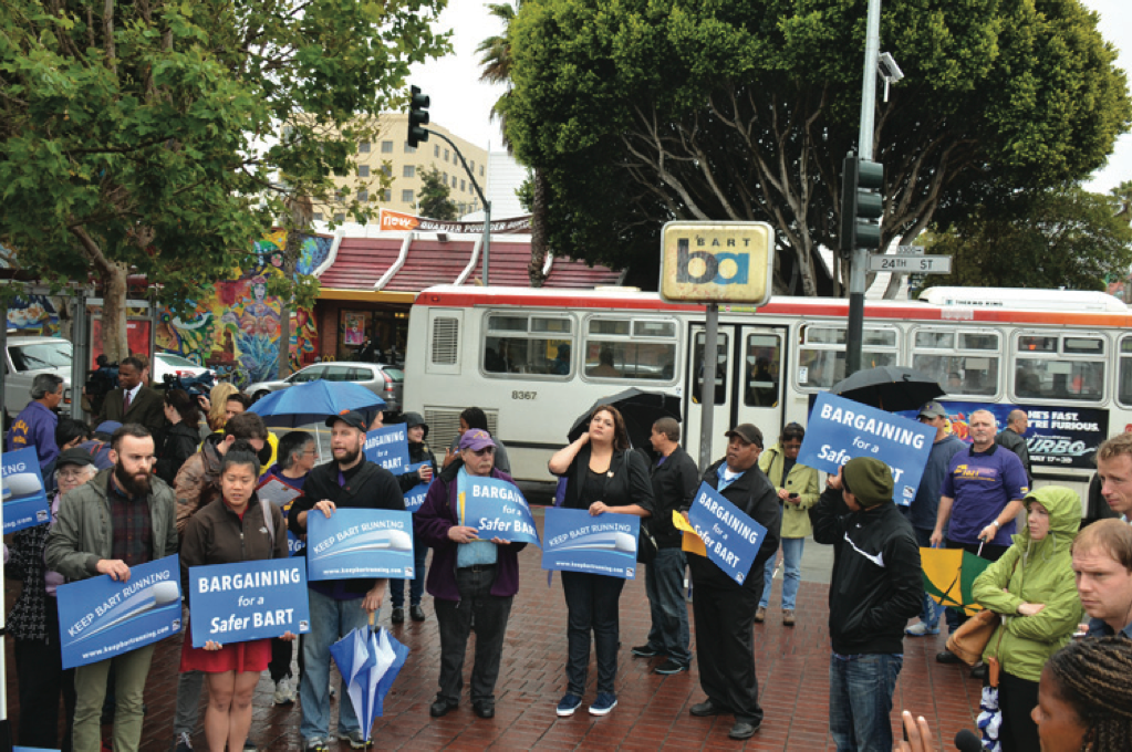 Protesters in front of a bus holding signs that say "Bargaining for a safer BART."