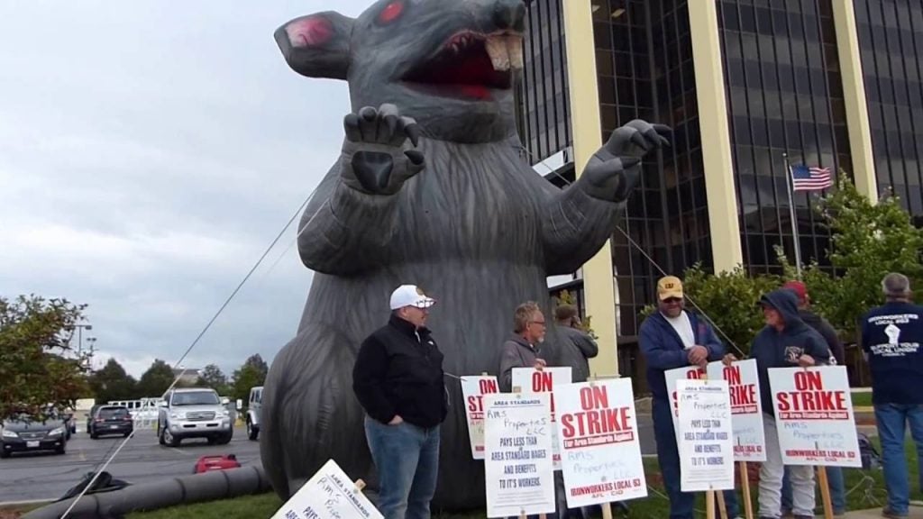 Protesters in front of a blow-up rat holding signs that say 