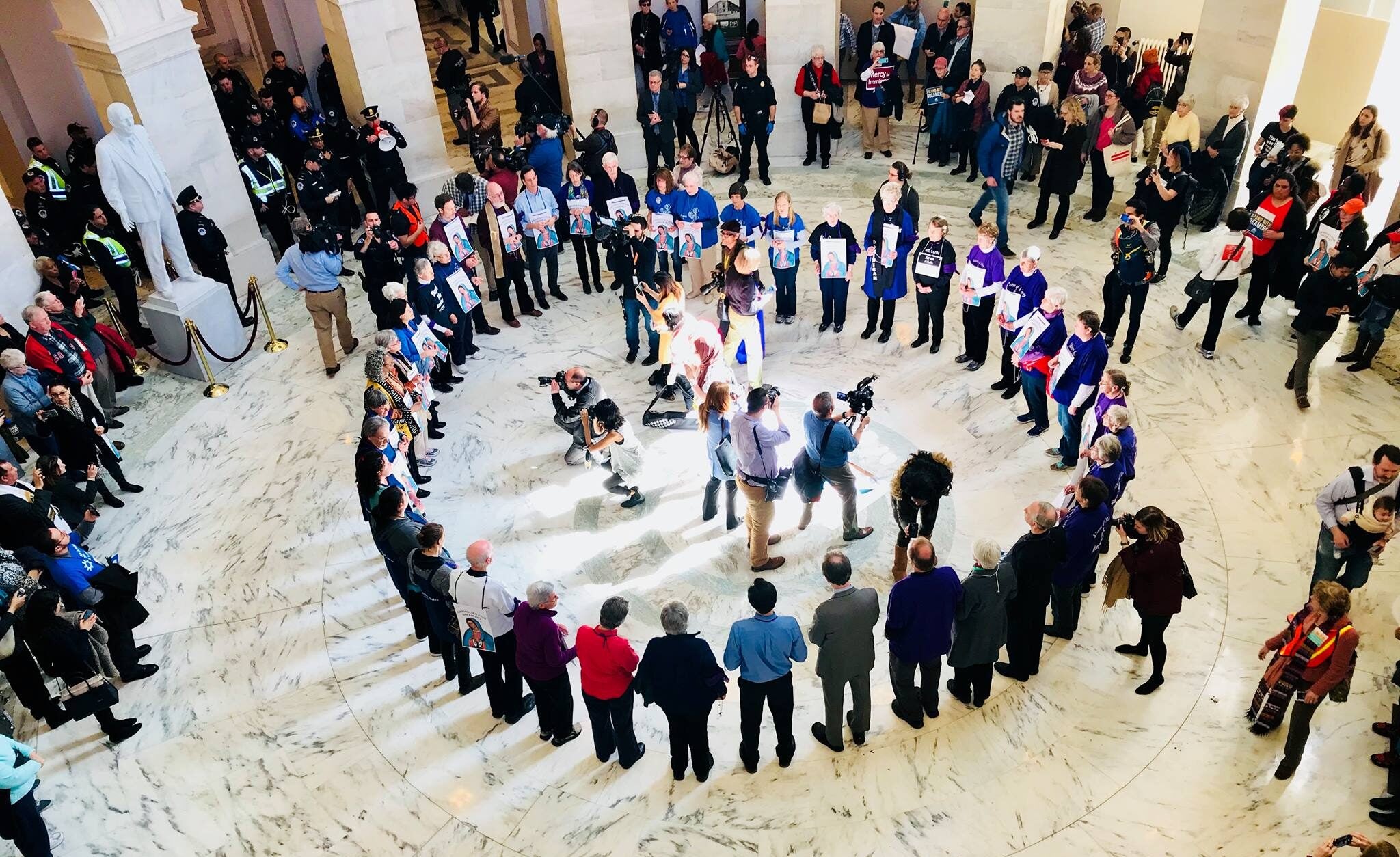 Faith protesters stand in a circle in a government building. Photographers take pictures from inside the circle.