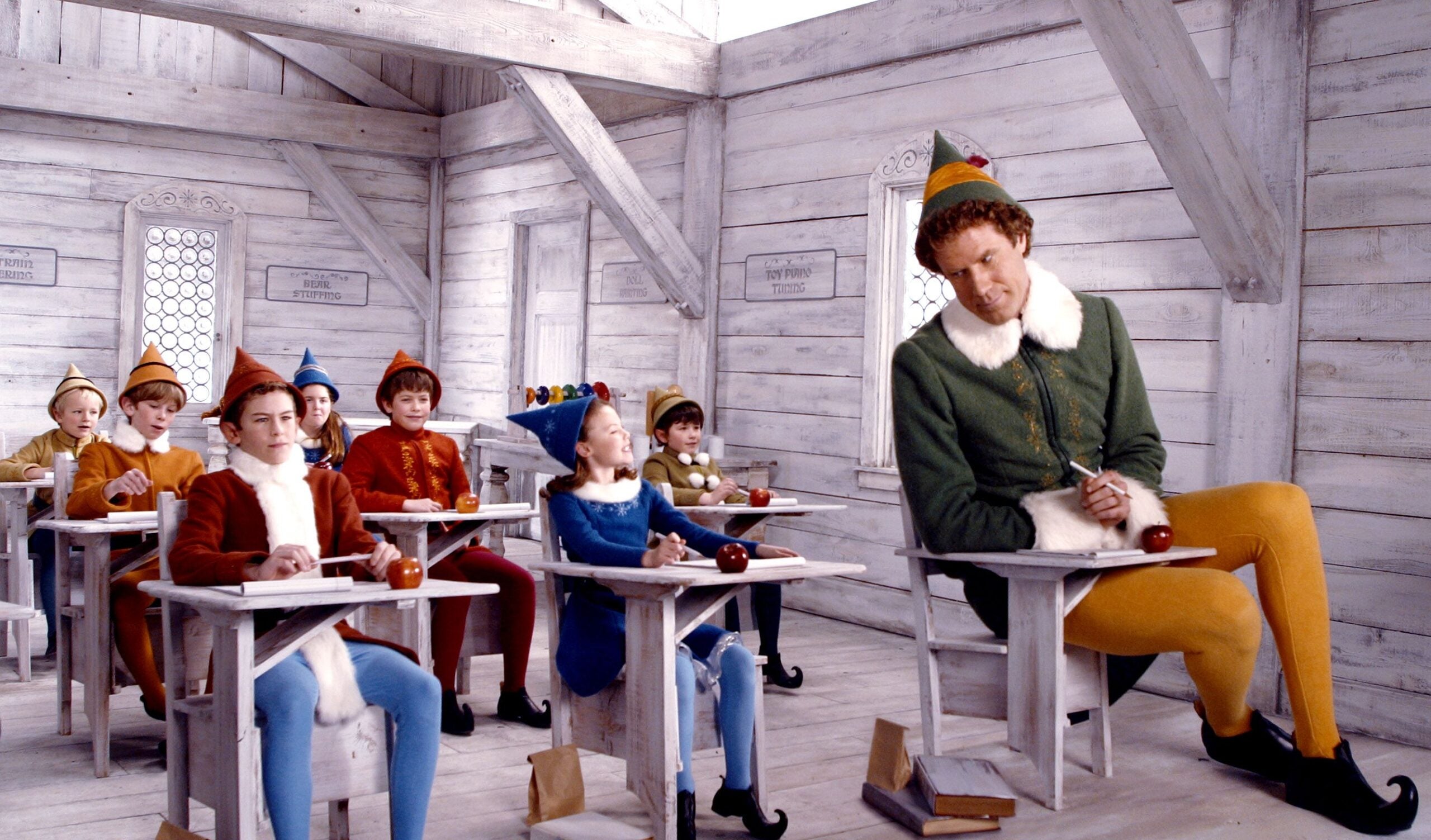 Still from the movie "Elf" in which the human-sized main character, Buddy, sits in a classroom with elf-sized children. All of the characters are dressed in elf attire.