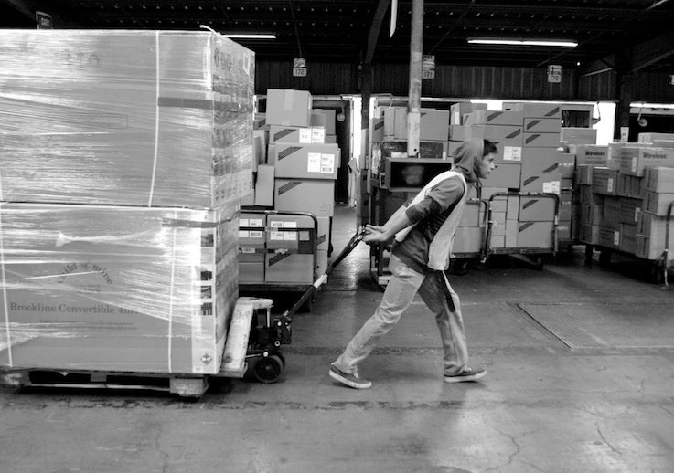 Black and white photo of a factor worker pulling a stack of boxes on a dolly.