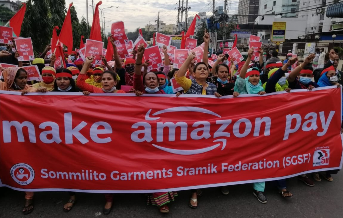 Protesters with raised fists hold a red banner that reads "make amazon pay."