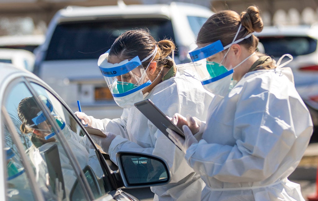 Medical professionals wearing gowns, gloves, face masks, and face shields stand in front of a car window. One is holding an iPad and the other is writing something down on a clipboard.
