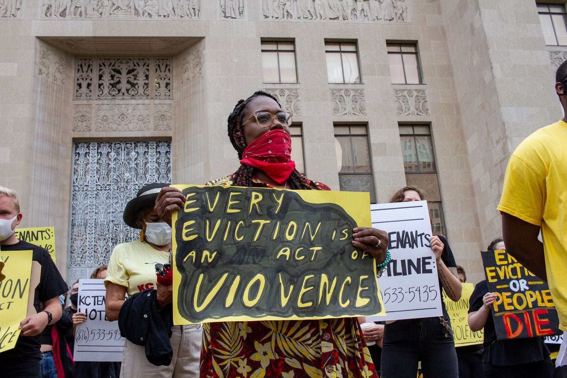 Protesters holding signs advocating for tenant rights. In the front, a person wearing a red bandana as a mask holds a sign with the words 