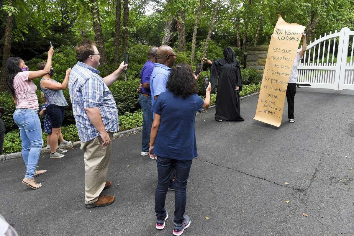 Person stands on the driveway of a house with a white gate holding a sign that reads "To: Warren Kanders. You owe reparations to the state of Connecticut for profiteering off of systemic racism and the suffering of others." A person wearing black robes and a black hood covering their face stands next to them. Several people are taking photos of the two protesters on their phones.