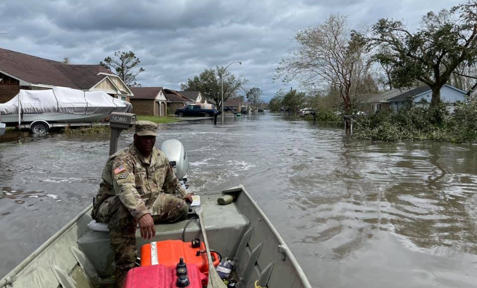 Man in a military uniform sails through a flooded neighborhood in a small boat.
