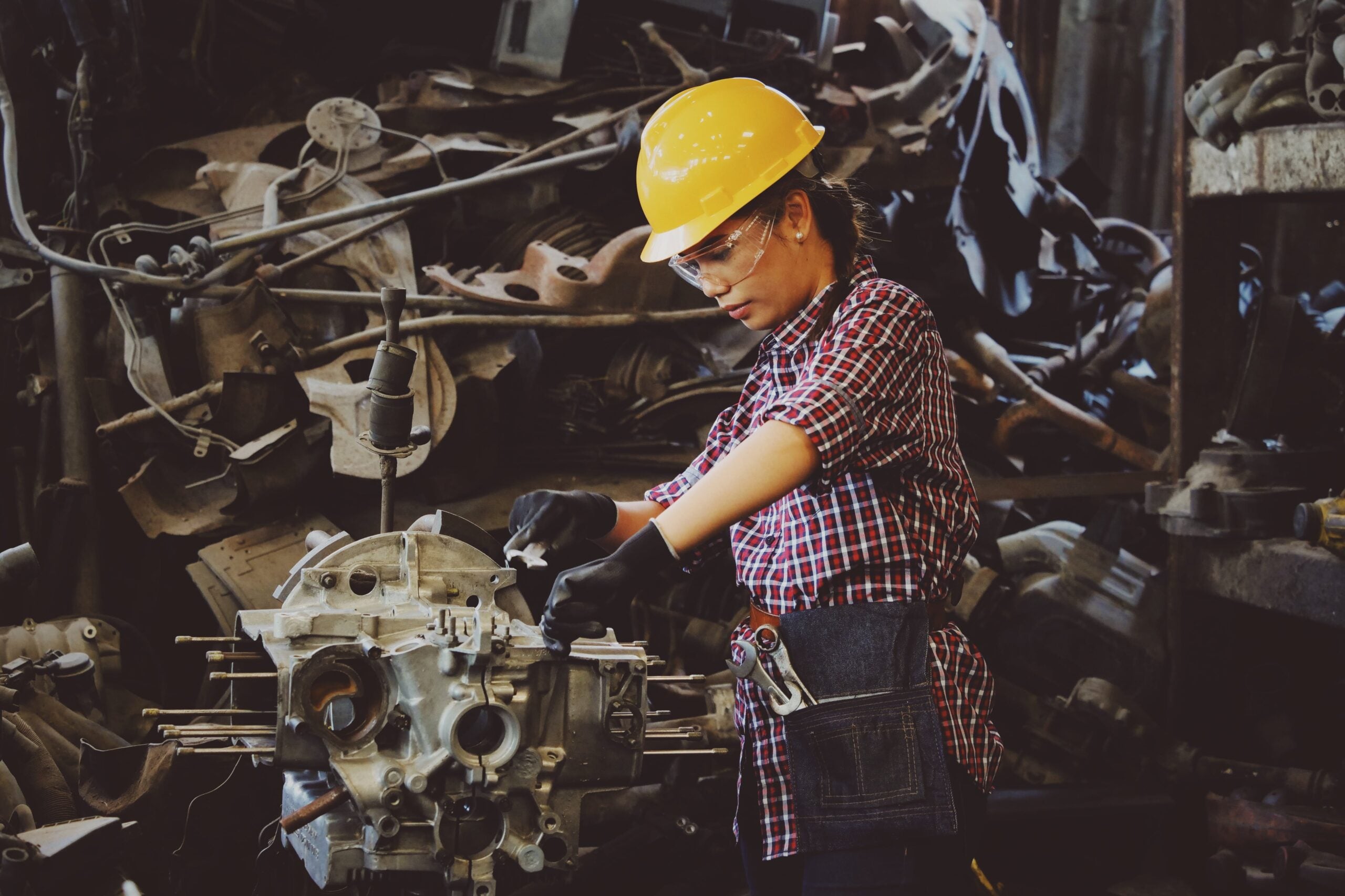 Woman in yellow hard hat and red checkered shirt working on mechanical parts in a factory